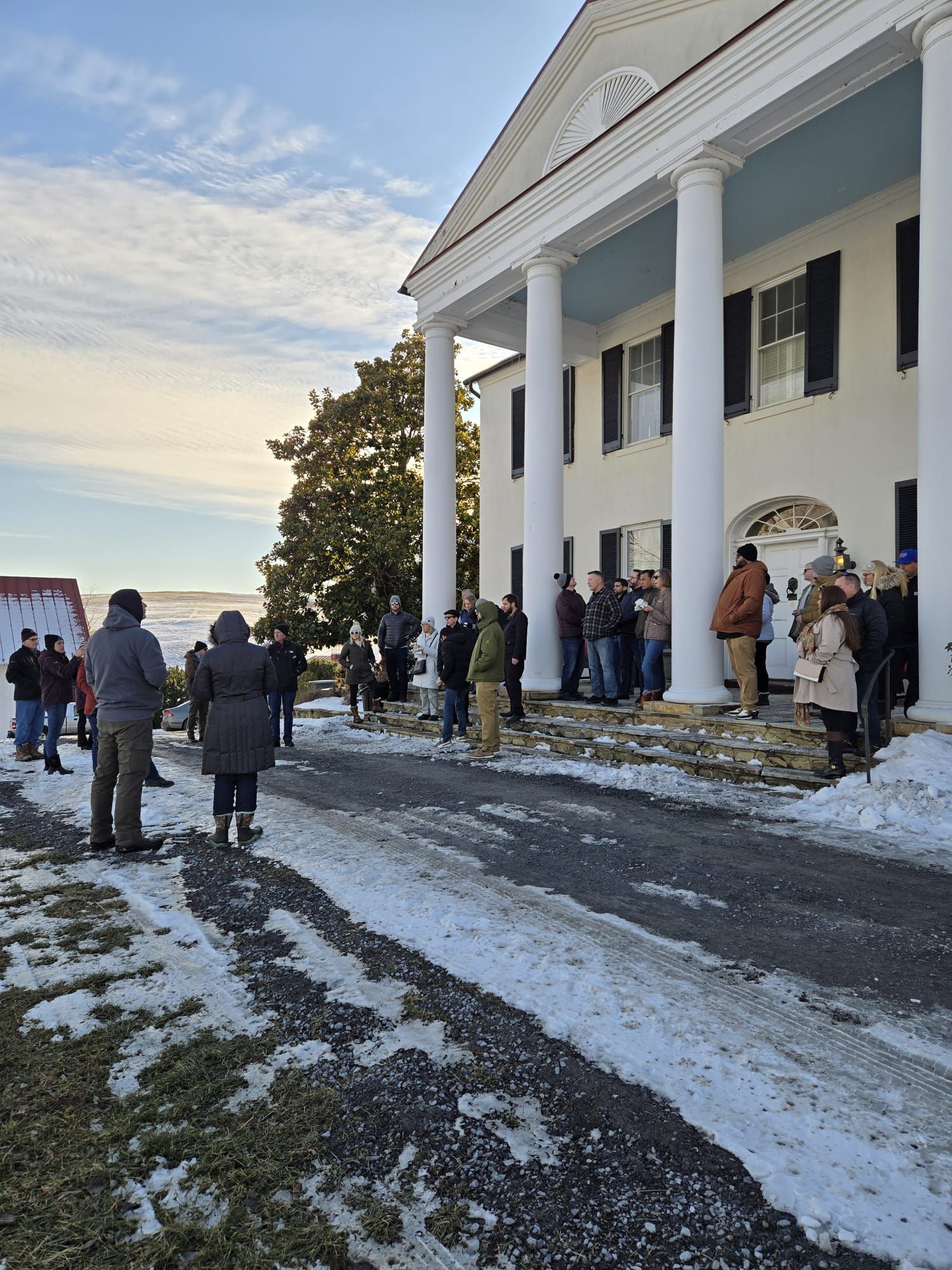 A bunch of people standing in a snowy driveway in front of a colonial house with pillars.